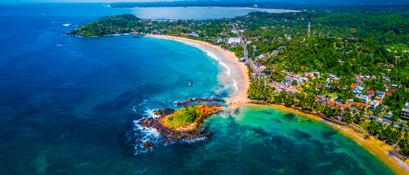 Mirissa's curved sandy beach and vibrant turquoise ocean with a lush green island near the shore.