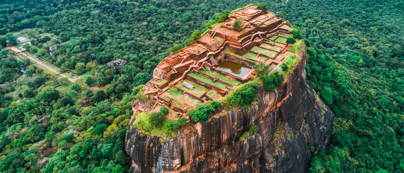 Ancient Sigiriya Rock Fortress with terraced gardens and ruins on its summit, surrounded by lush green forest.