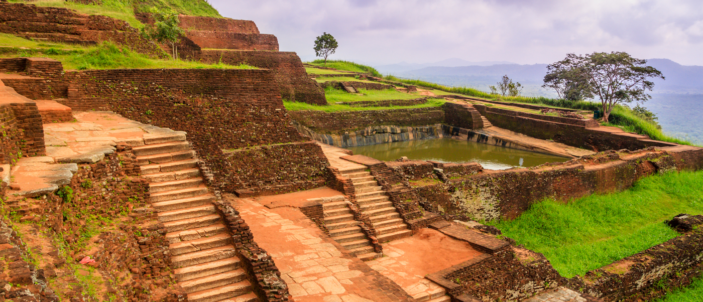 Ancient stone staircases and terraced ruins of Sigiriya Rock Fortress, with a serene water pool and distant landscape.