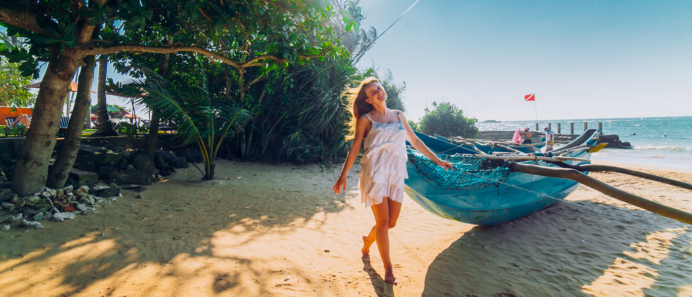A joyful woman strolls on the sandy Weligama beach beside a traditional blue fishing boat.