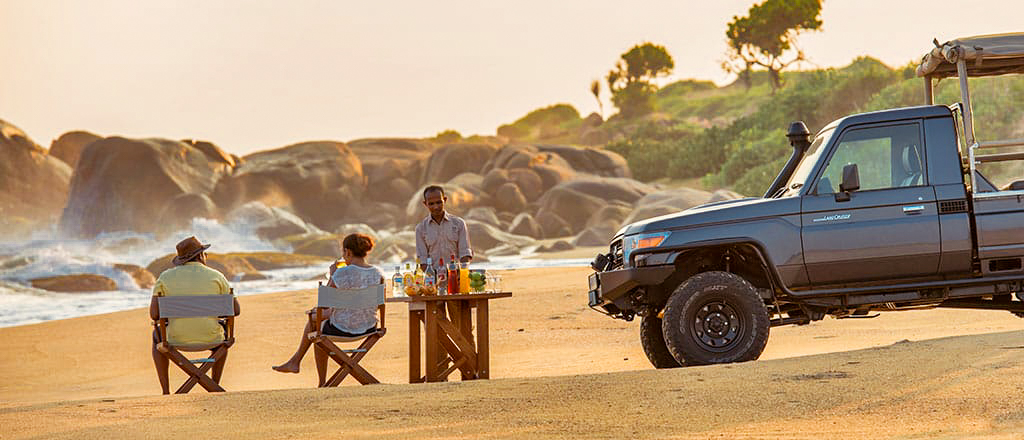 Guests enjoy a private beach sundowner with a safari vehicle and ocean waves at Yala National Park.