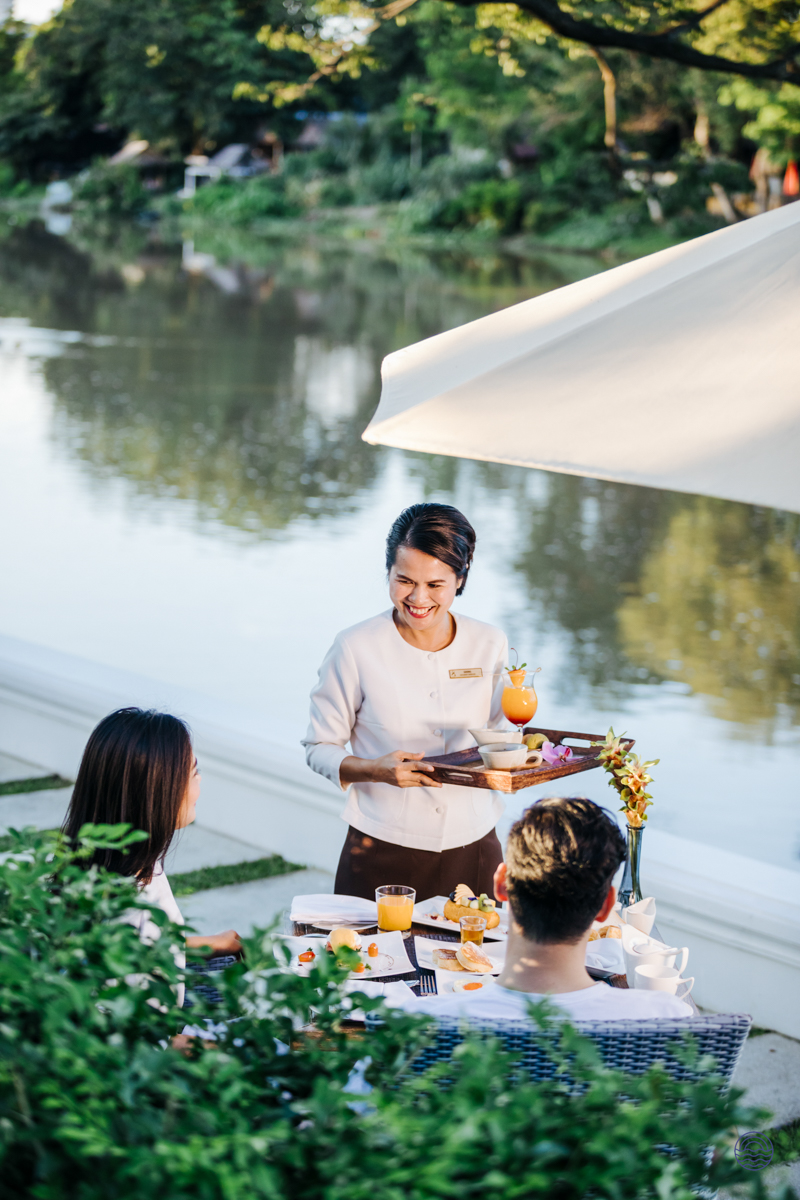 A smiling server delivers drinks to a couple enjoying a romantic riverside breakfast in Chiang Mai.