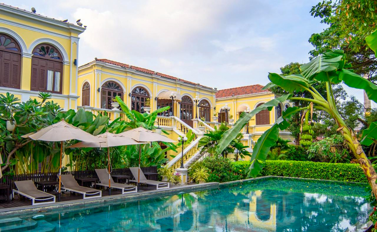 A beautiful yellow historic hotel building with a swimming pool and tropical gardens in Bangkok.