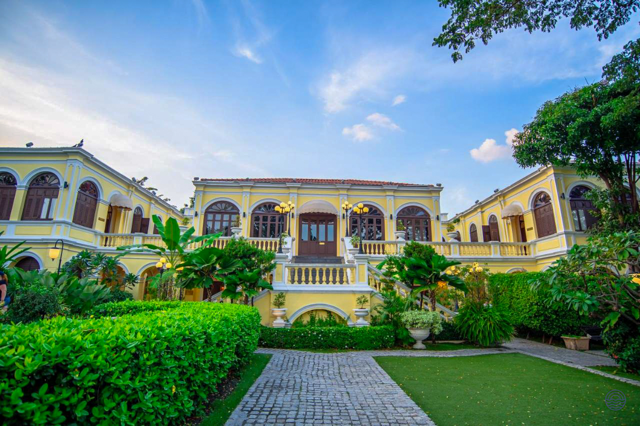 An elegant historic yellow mansion hotel with lush green gardens under a blue sky in Bangkok.