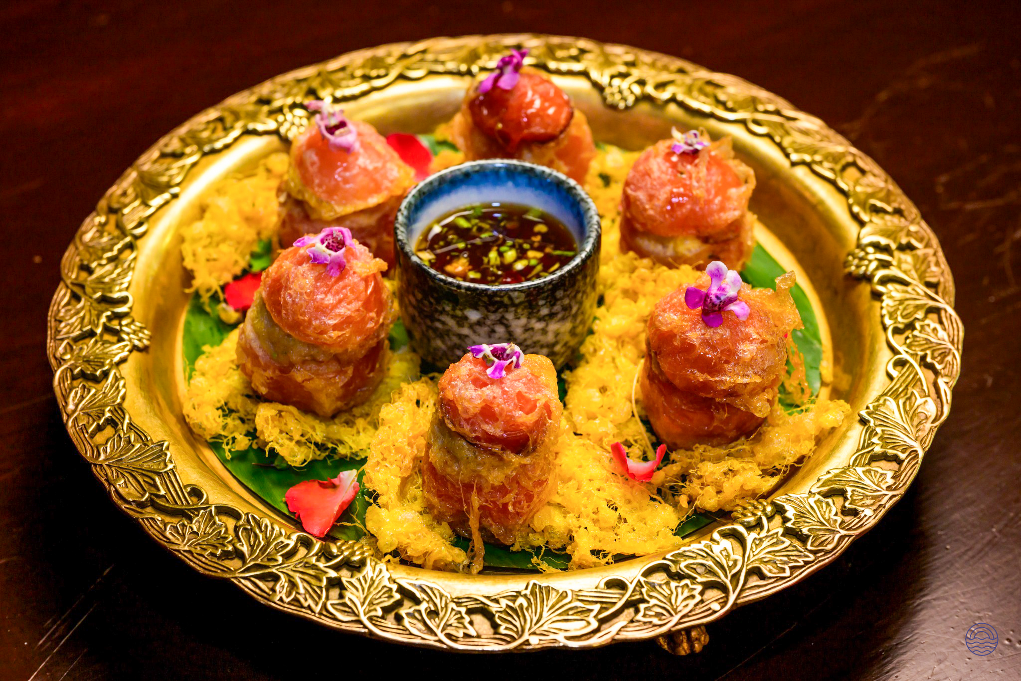 Six elegant Thai appetizers with purple flowers and dipping sauce on an ornate golden platter.