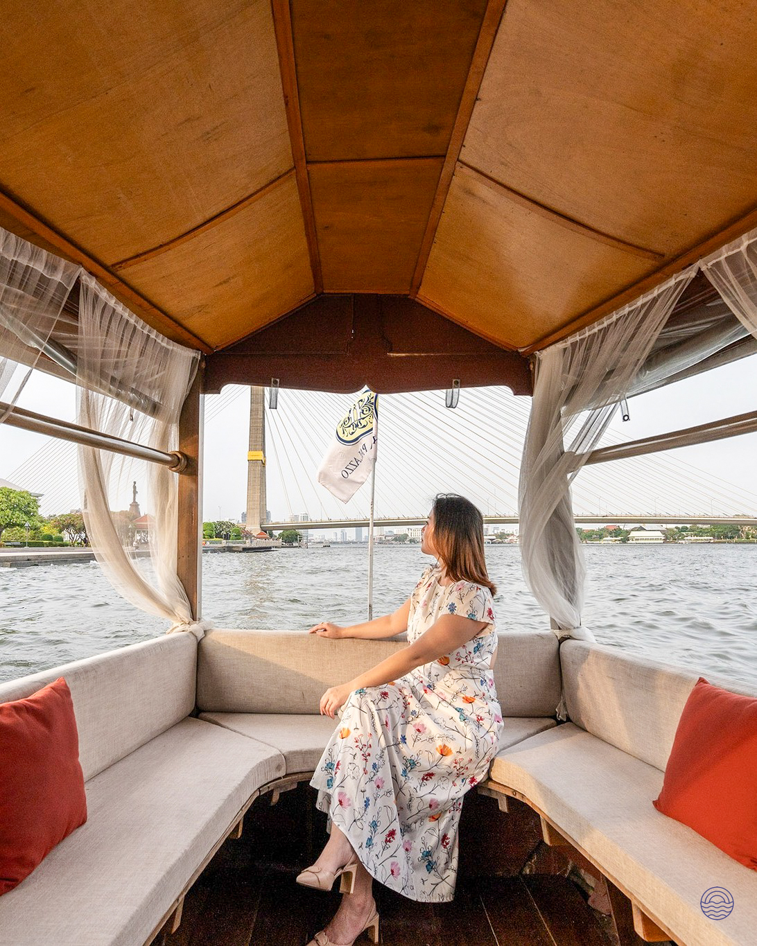 Woman in a floral dress relaxing on a traditional wooden boat, looking at the Chao Phraya River in Bangkok.