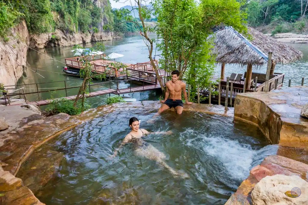 A couple relaxes in a natural hot spring overlooking a serene river in Kanchanaburi, Thailand.