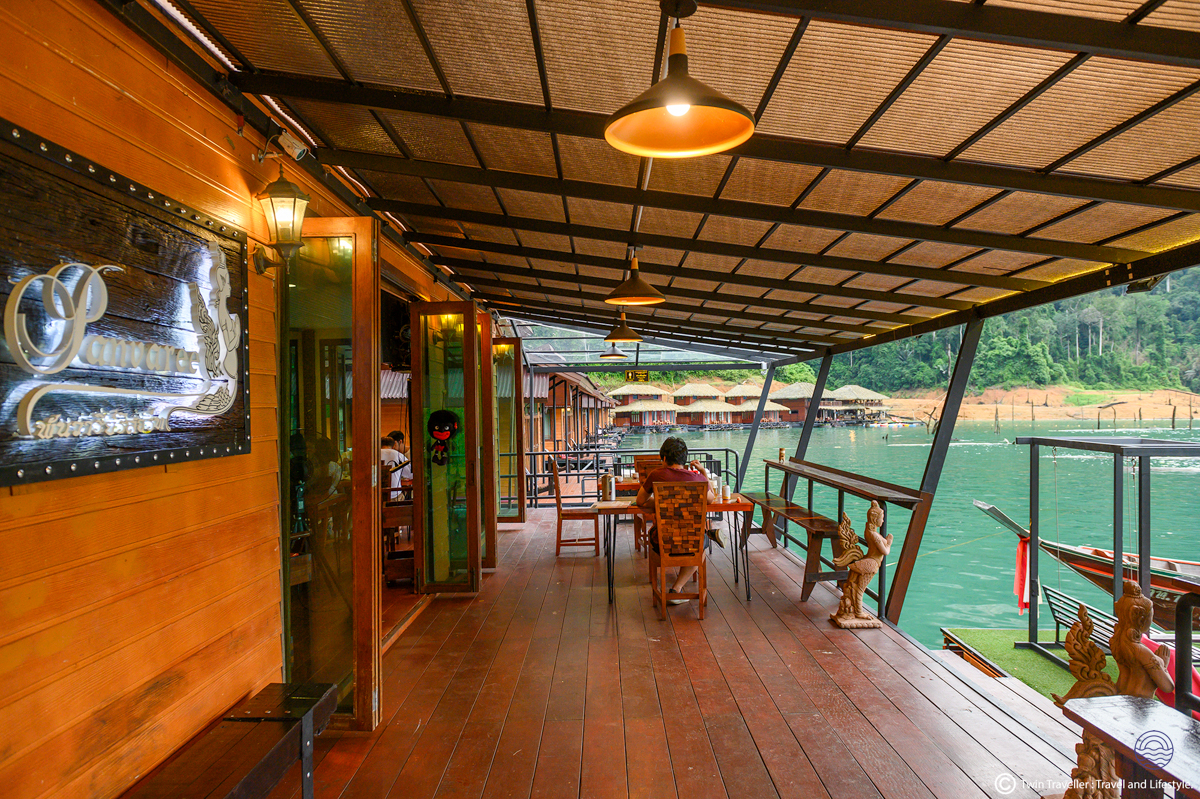 A person relaxes on the wooden deck of a floating resort, overlooking Khao Sok's calm green waters.