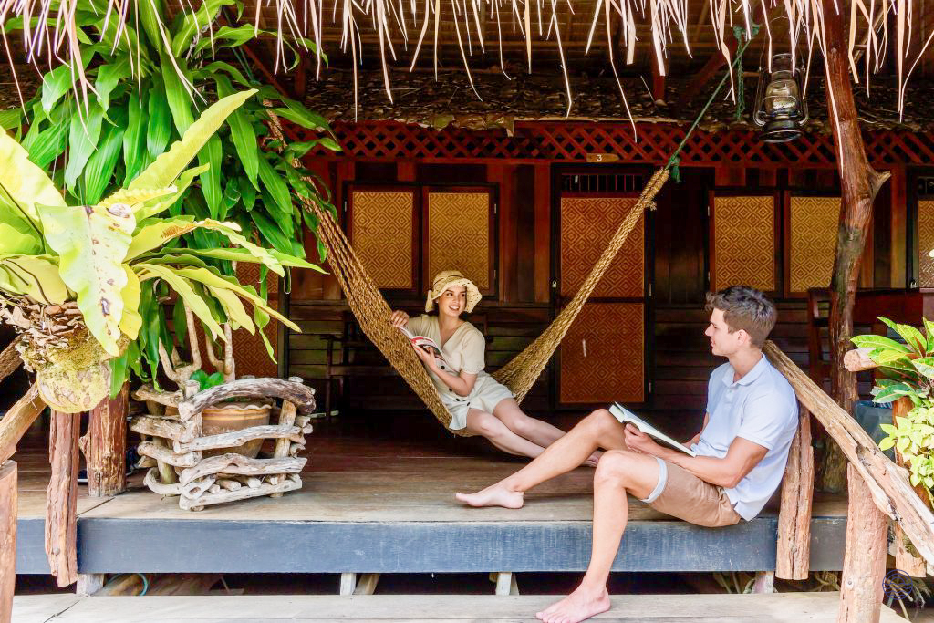A couple relaxes, reading books on the wooden porch of their River Kwai jungle bungalow.