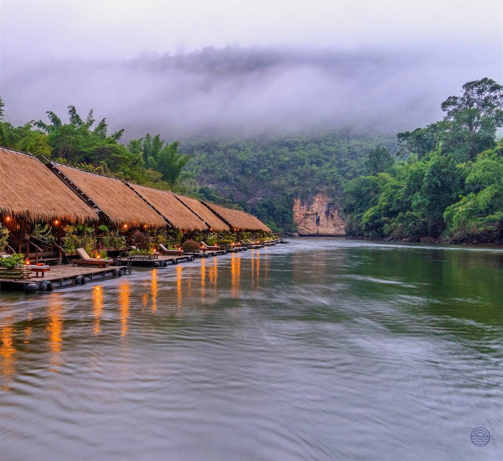 Thatched-roof bungalows float on the misty River Kwai, surrounded by lush jungle in Kanchanaburi, Thailand.