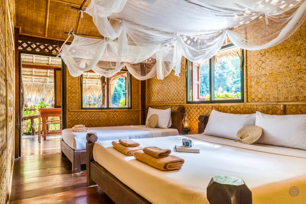 Inviting bungalow room in Kanchanaburi with bamboo walls, two beds, and mosquito nets overlooking the jungle.