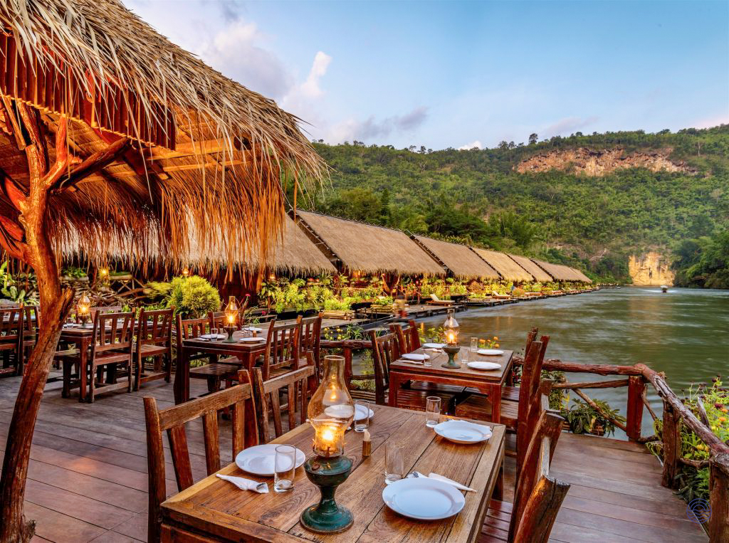 Peaceful riverside restaurant with thatched roofs and wooden tables overlooking the River Kwai in Kanchanaburi.