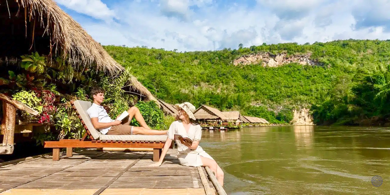 A couple reads and relaxes on their River Kwai jungle raft bungalow deck, surrounded by jungle.