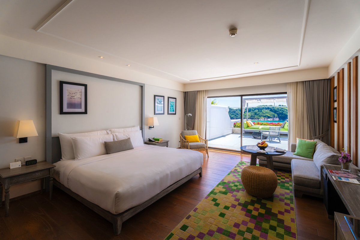 Luxurious hotel room with a king bed, lounge area, and a balcony view of Phuket's green hills.