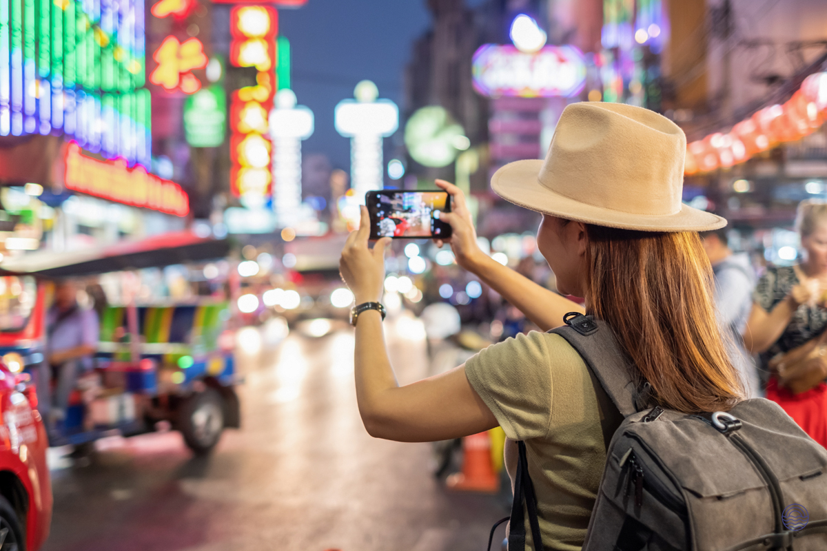 A woman in a hat photographs the vibrant, neon-lit streets of Bangkok at night.