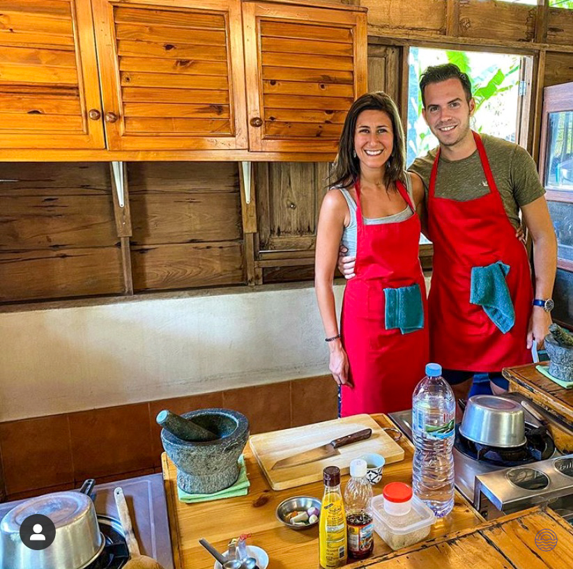 Smiling couple in red aprons enjoying a hands-on Thai cooking class in Chiang Mai.