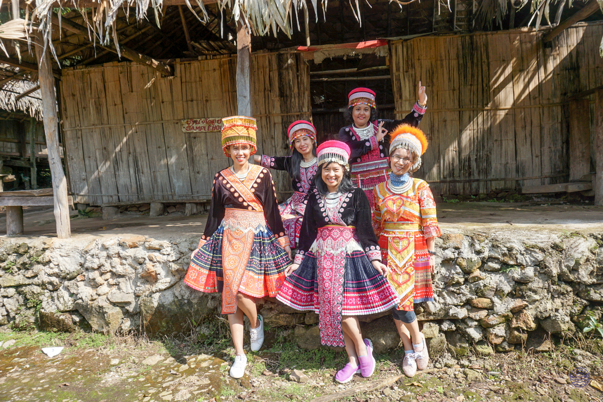 Five smiling women in traditional Hmong attire pose in front of a rustic village house in Chiang Mai.