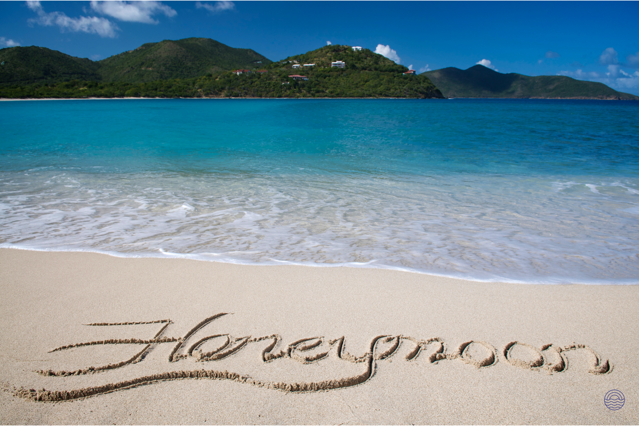 Honeymoon written in the sand on a pristine beach with turquoise water and green hills.