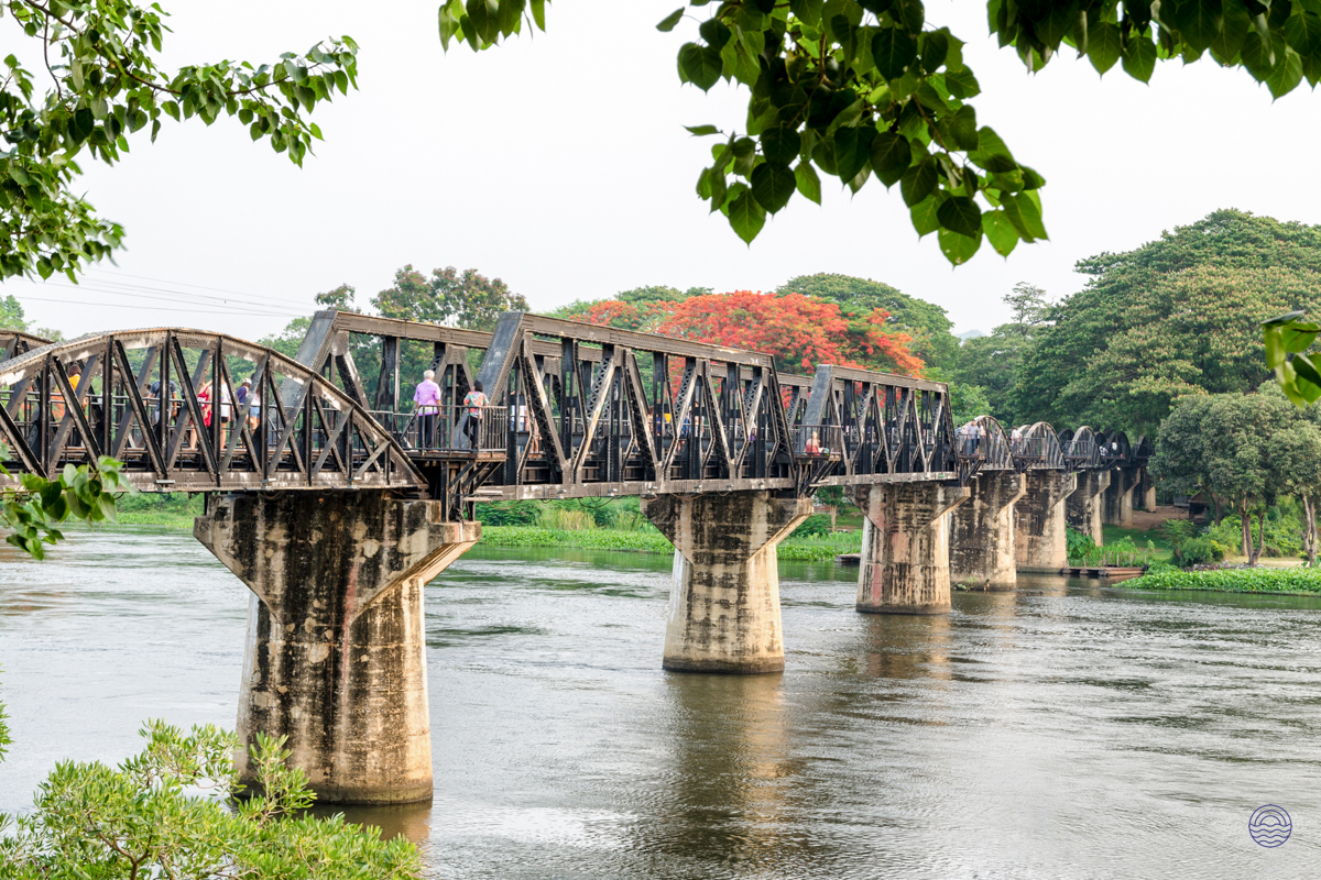 Visitors stroll across the historic Bridge on the River Kwai in Kanchanaburi, surrounded by lush tropical trees.