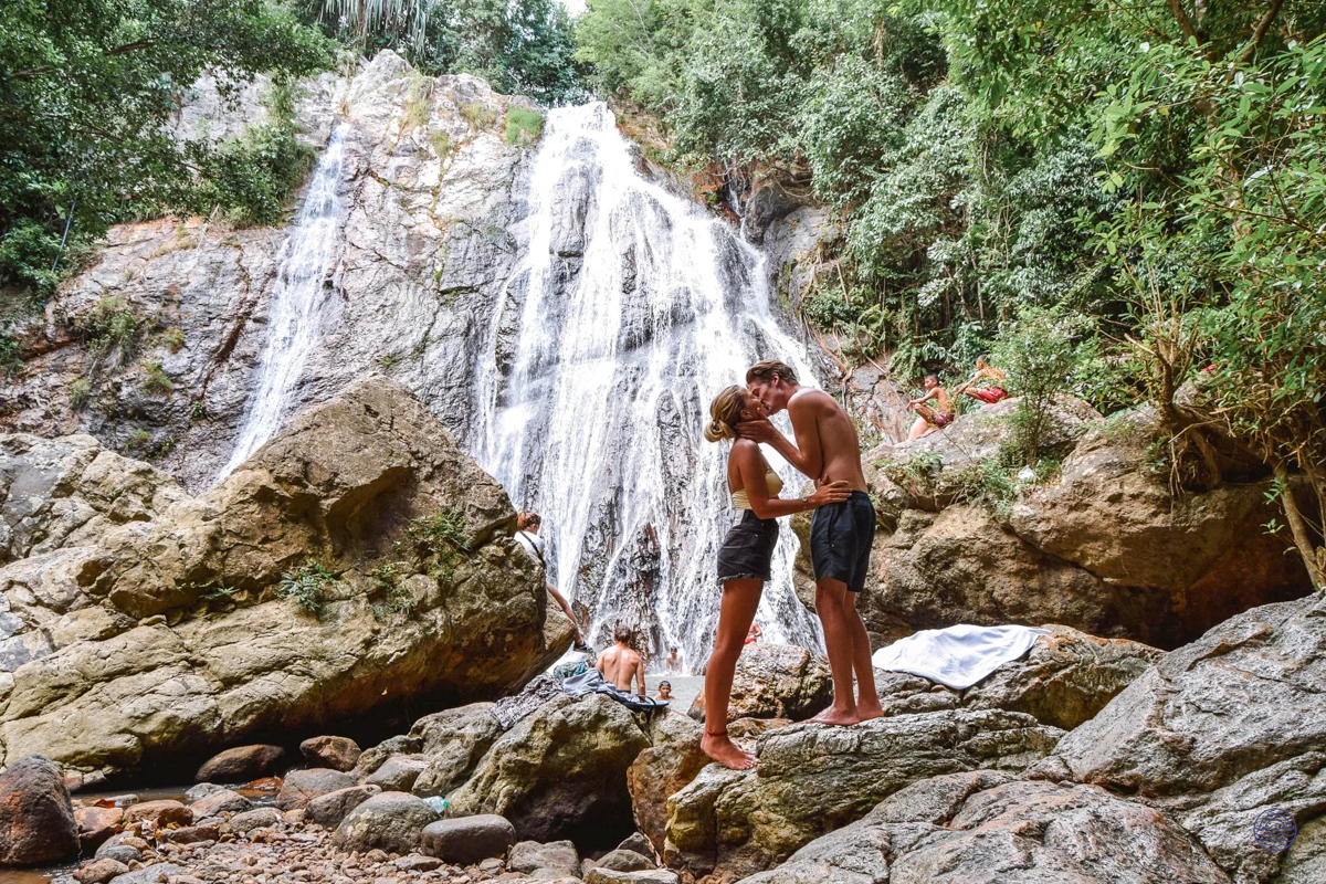 A couple shares a romantic kiss on rocks in front of a magnificent waterfall in Koh Samui.