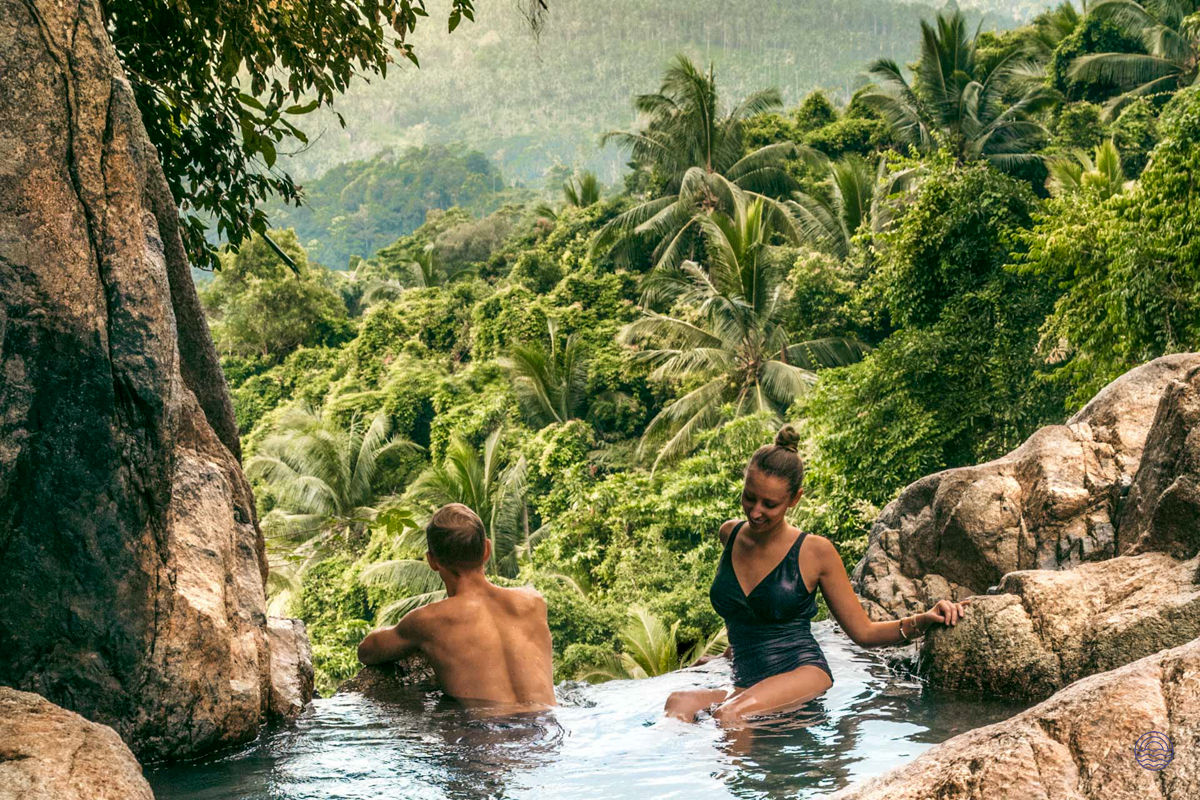 A couple relaxes in a natural pool with lush tropical jungle views on Koh Samui.