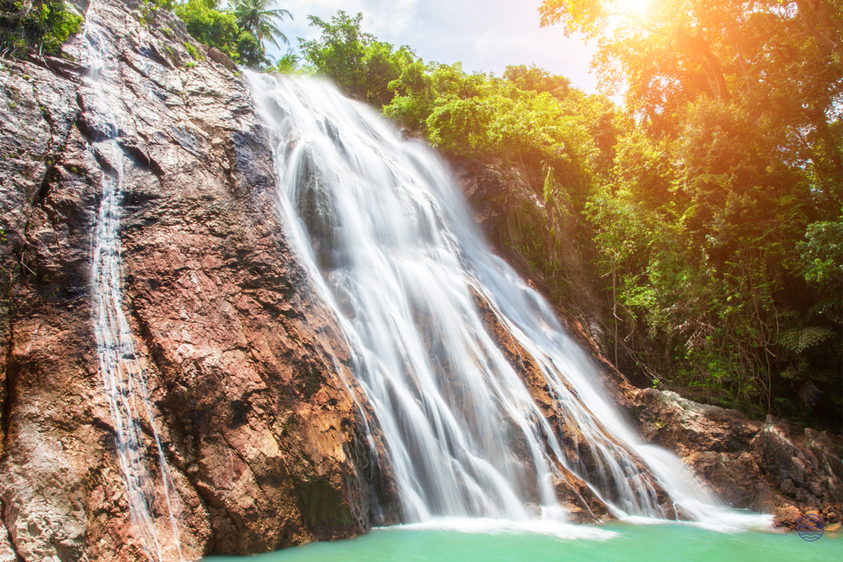 A stunning waterfall cascades over reddish-brown rocks into a vibrant turquoise pool on Koh Samui.