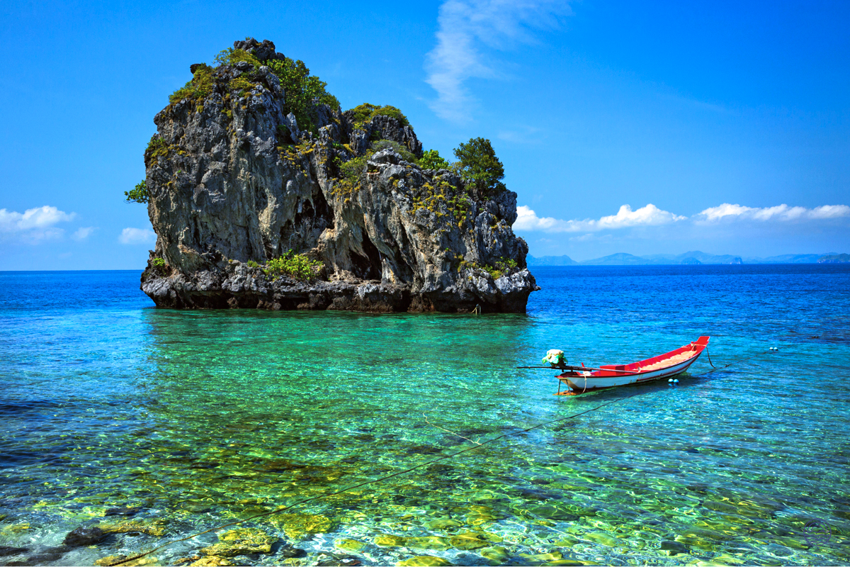 A red longtail boat floats in the clear turquoise waters of Krabi, near a beautiful limestone island.