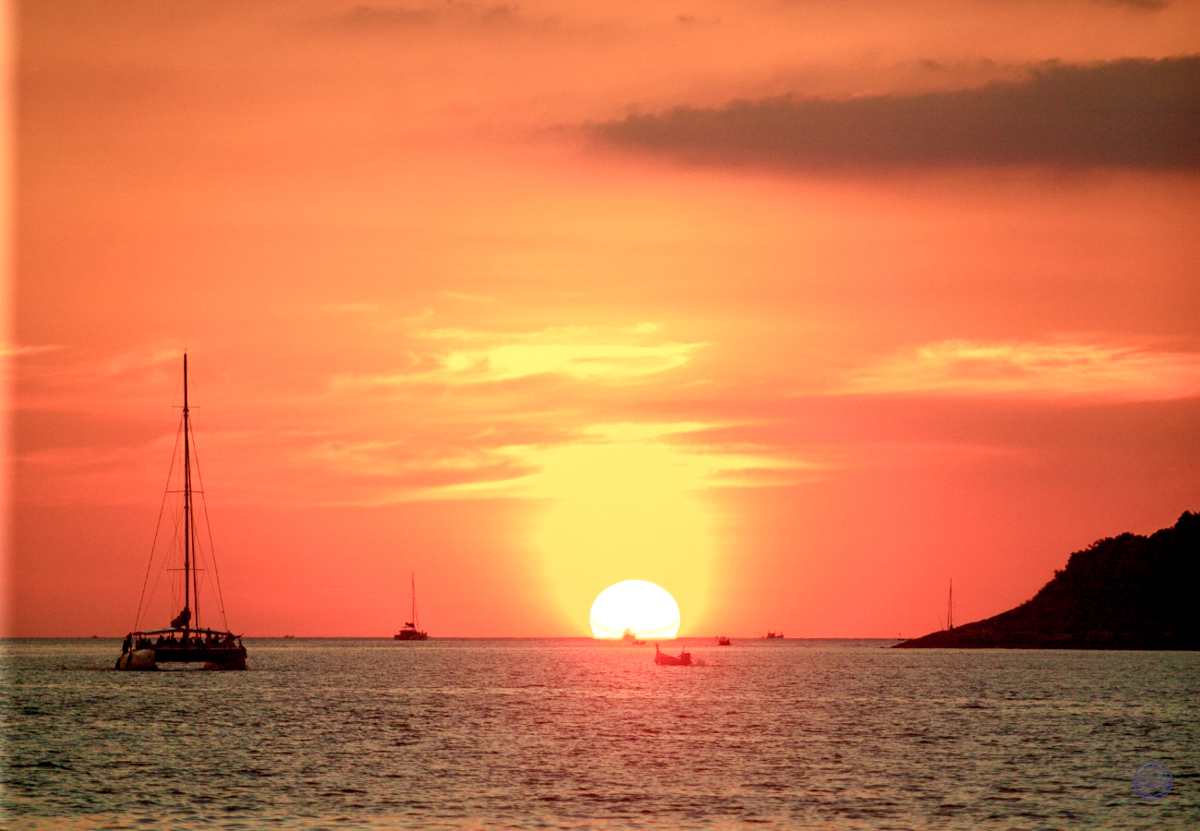 A vibrant orange sunset over the Andaman Sea, with boats cruising near the Phuket coastline.