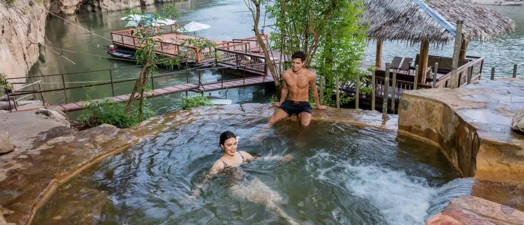 A couple enjoys a natural rock pool with a small waterfall overlooking a river and cliffs in Thailand.
