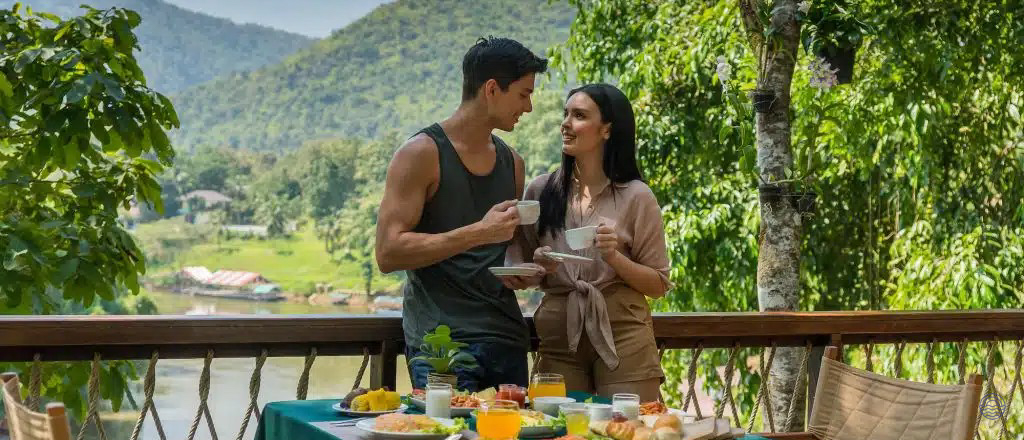 A happy couple enjoys breakfast and coffee on a balcony overlooking serene mountains and a river in Chiang Mai.