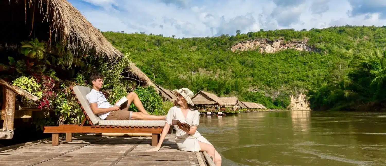 A couple relaxes on a wooden deck by the River Kwai, reading books amidst jungle rafts and green hills.
