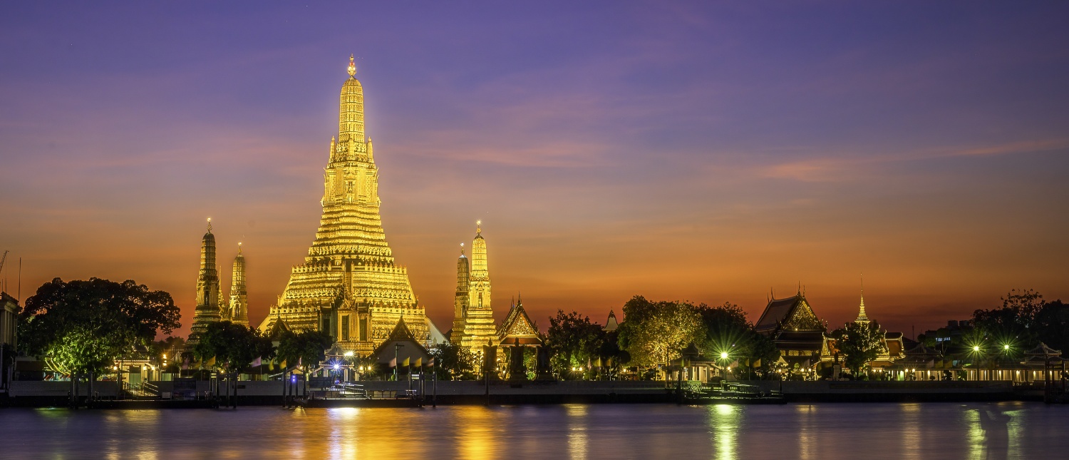 The majestic Wat Arun temple glows golden at twilight, reflected beautifully on the river in Bangkok.