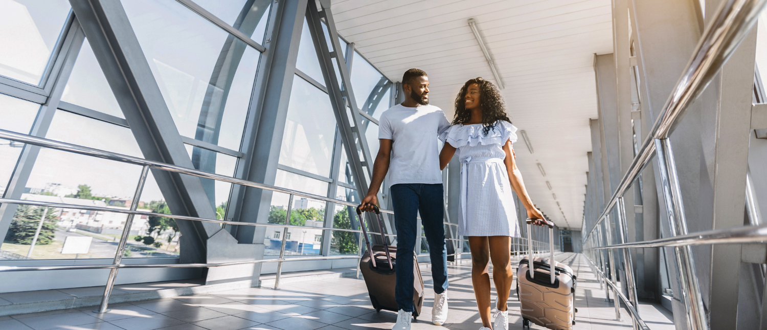 A smiling couple walks side-by-side through a bright airport walkway, pulling their luggage.