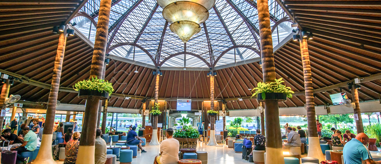 Travelers relax in the open-air Koh Samui Airport terminal, featuring tropical decor and a high glass roof.