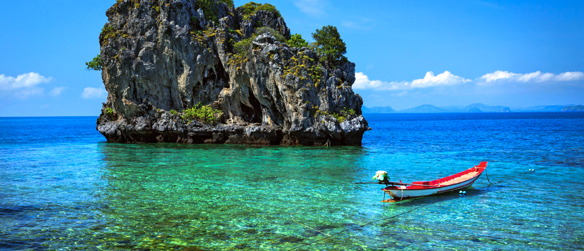 A small red longtail boat floats on clear turquoise water near a towering limestone island in Krabi.