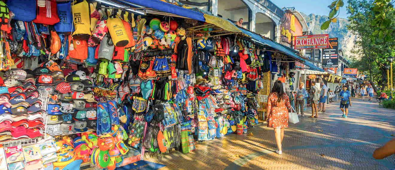 People browse vibrant market stalls overflowing with colorful clothes and souvenirs on a sunny street in Krabi.