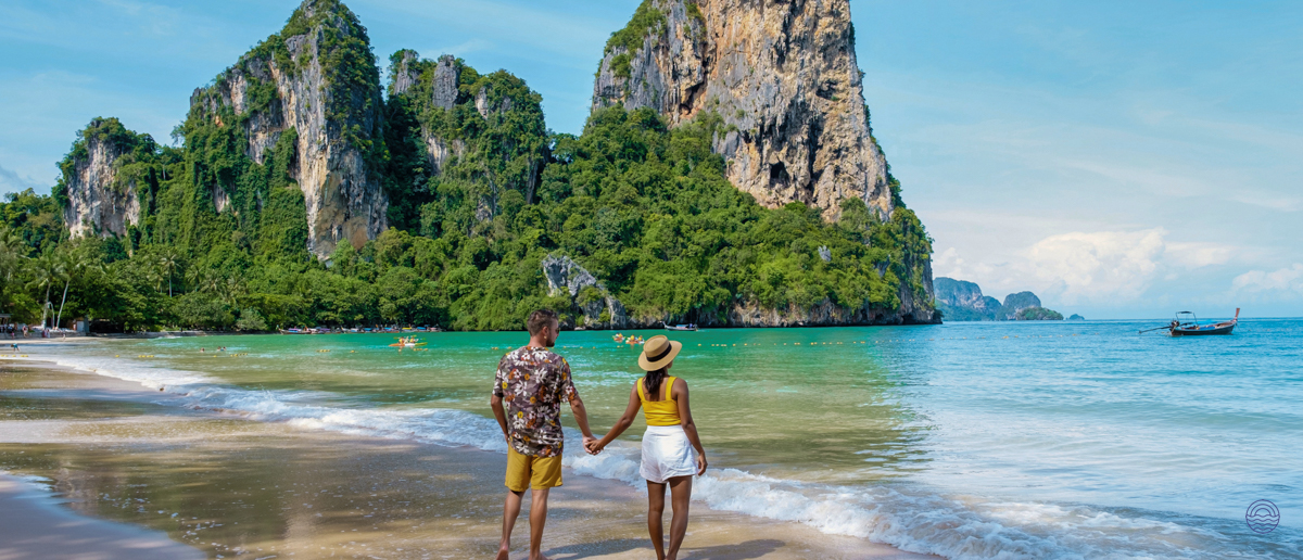A couple holds hands walking on a sandy beach towards the turquoise sea, dramatic limestone karsts in Krabi.
