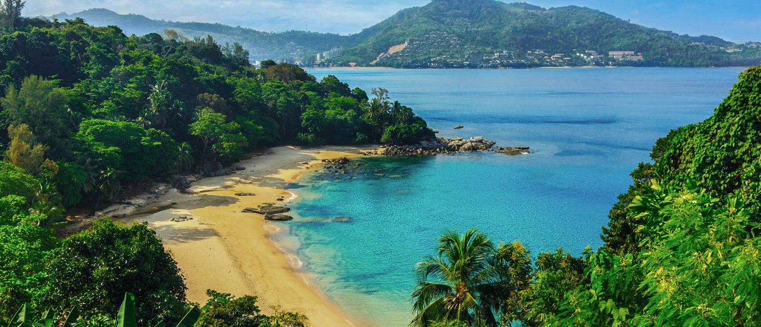 Lush green hills surround a beautiful golden beach and clear turquoise bay in Phuket.
