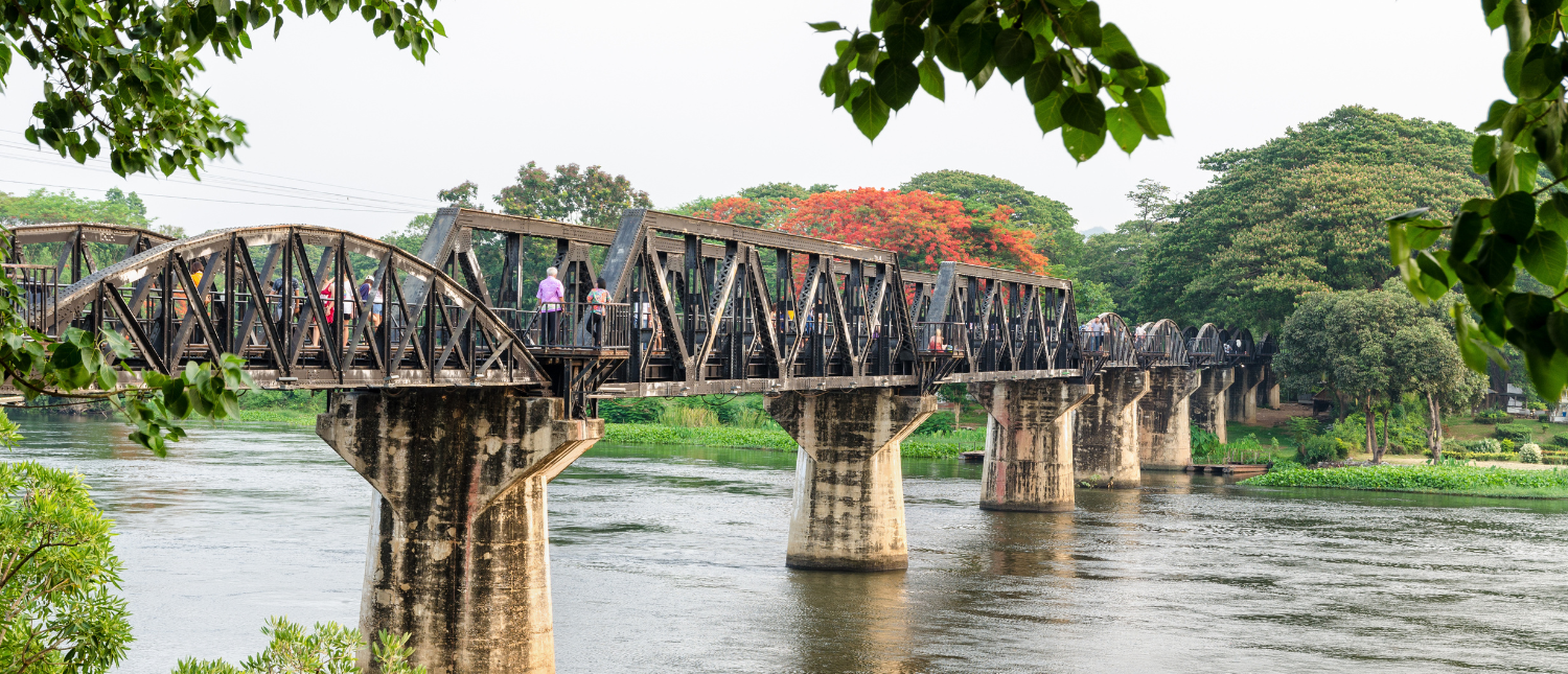 Visitors explore the historic River Kwai Bridge in Kanchanaburi, crossing a calm river under a cloudy sky.