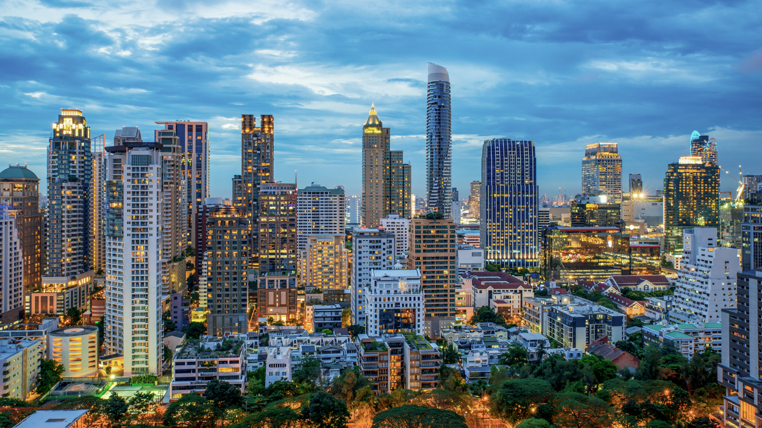 A panoramic view of the Bangkok skyline at dusk, with numerous illuminated skyscrapers under a cloudy sky.