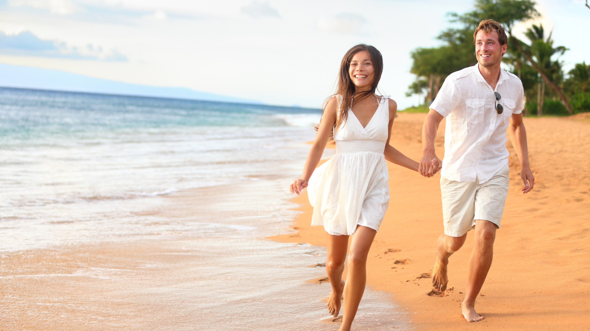 Smiling couple running barefoot on a golden sand beach next to the ocean waves.