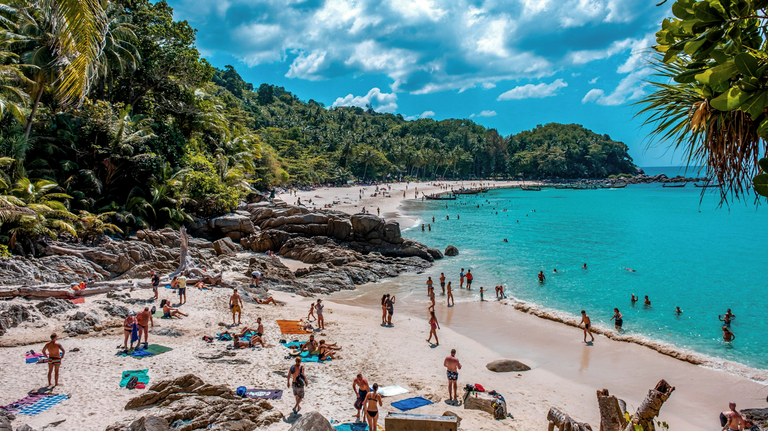 Many people swim and sunbathe on a vibrant Phuket beach with turquoise water and lush hills.