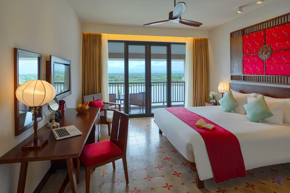 Comfortable hotel room in Hoi An with a large bed, desk, and balcony overlooking a river.