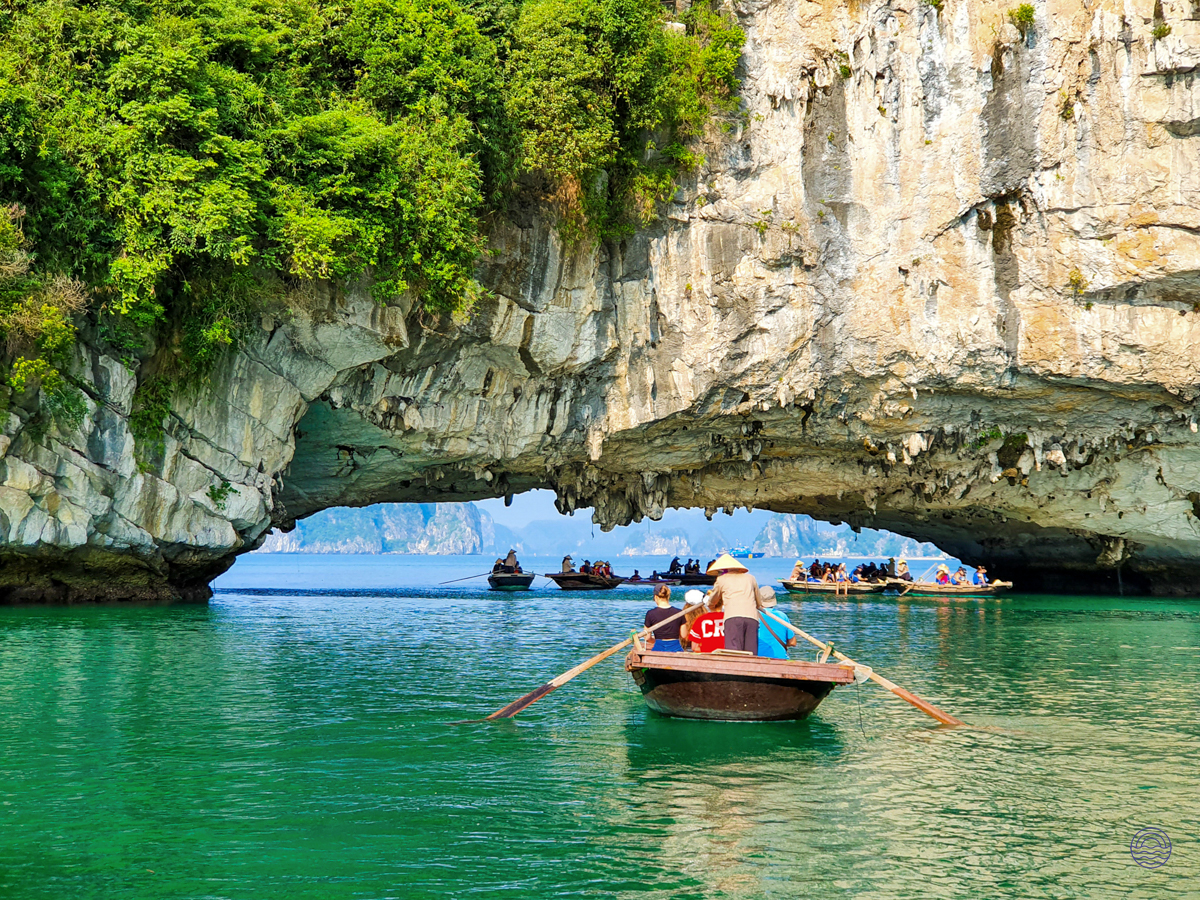 A person in a conical hat rows a traditional boat through a stunning rock arch in Ha Long Bay.