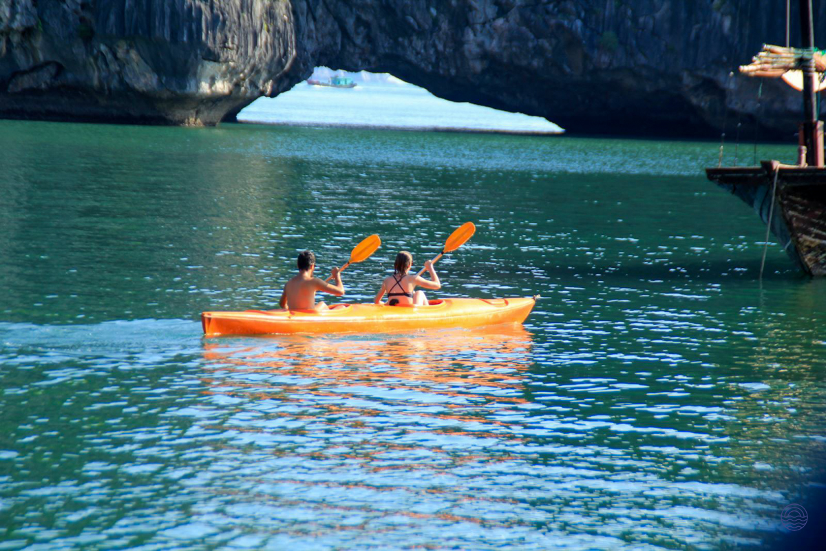 Two people kayak through the serene waters of Halong Bay, passing a natural rock archway.