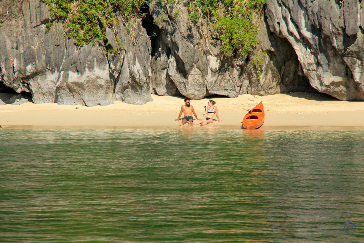 A couple relaxes on the pristine sands of Ba Trai Dao Beach, Halong Bay, beside a kayak.