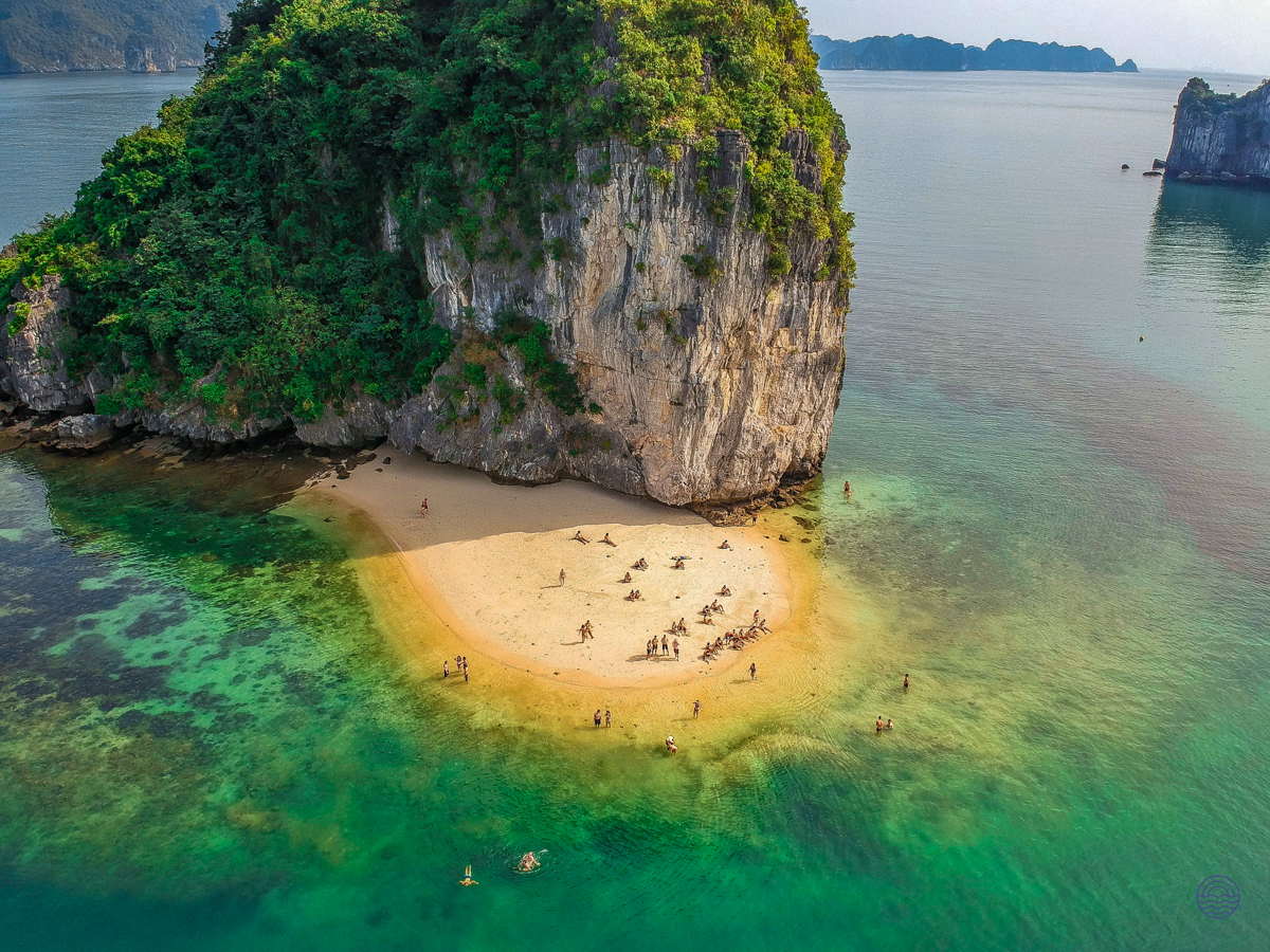 Visitors swim and relax on the serene sandy beach of a lush karst island in Halong Bay.