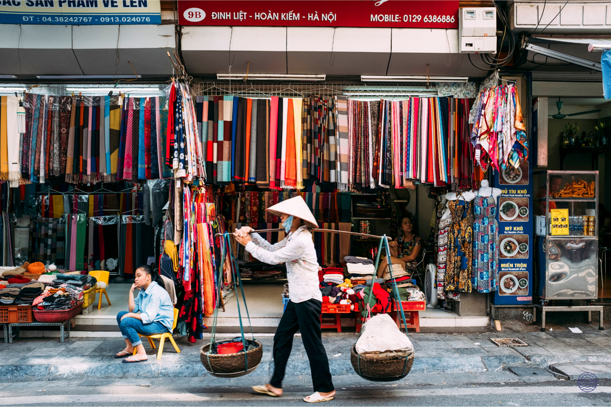 A woman in a conical hat carries baskets on a shoulder pole past a vibrant fabric shop in Hanoi.