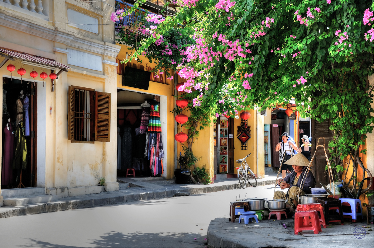 A charming street scene in Hoi An with a vendor, yellow buildings, and vibrant pink bougainvillea.