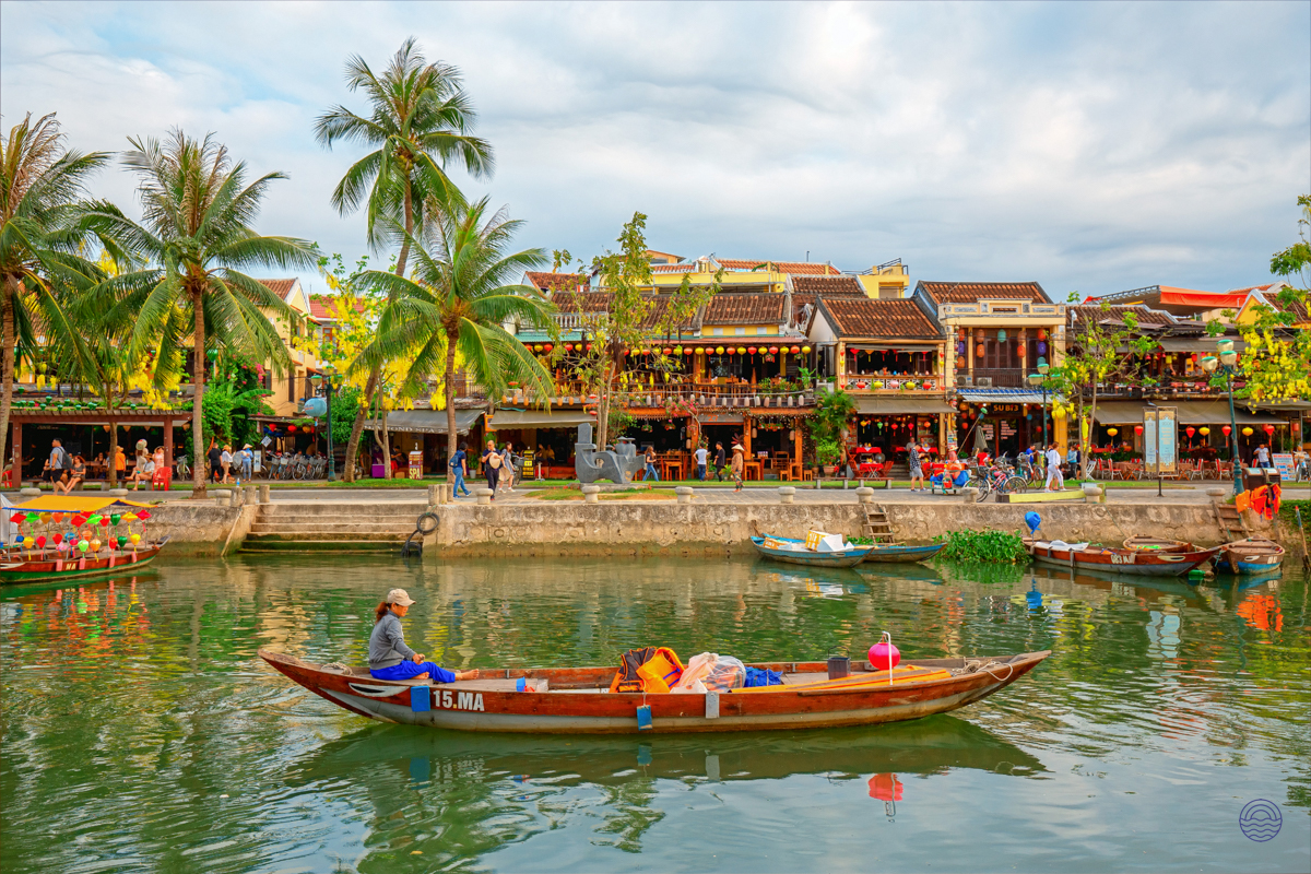 A person paddles a wooden boat on a river lined with historic buildings and palm trees in Hoi An.